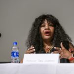 A woman with long hair and a necklace is speaking at a table.